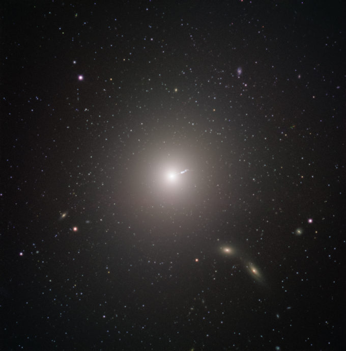 Messier 87 (M87) es una enorme galaxia elíptica ubicada a unos 55 millones de años luz de la Tierra, visible en la constelación de Virgo. Fue descubierta por Charles Messier en 1781, pero no fue identificado como una galaxia hasta el siglo XX. Se encuentra entre las galaxias más grandes del universo local: posee el doble de masa de nuestra propia galaxia, la Vía Láctea, y contiene hasta diez veces más estrellas. Además de su tamaño original, M87 tiene algunas características muy únicas. Por ejemplo, contiene un número inusualmente alto de cúmulos globulares: mientras nuestra Vía Láctea contiene menos de 200, M87 tiene aproximadamente 12.000, que según algunos científicos teorizan habría recolectado de sus vecinos más pequeños. Al igual que con todas las demás galaxias grandes, M87 tiene un agujero negro supermasivo en su centro. La masa del agujero negro en el centro de una galaxia está relacionada con la masa de la galaxia en general, por lo que no debería sorprender que el agujero negro de M87 sea uno de los más masivos conocidos. El agujero negro también puede explicar una de las características más energéticas de la galaxia: un chorro de relativista de materia que se expulsa a casi la velocidad de la luz. El agujero negro fue objeto de observaciones para cambiar paradigmas por parte del Event Horizon Telescope. El EHT lo eligió como el objetivo de sus observaciones por dos razones. Si bien la resolución de la EHT es increíble, también tiene sus límites. En vista que los agujeros negros más masivos son también más grandes en diámetro, el agujero negro central de M87 presentaba un objetivo inusualmente grande, lo que significa que se podría obtener imágenes más fácilmente que los agujeros negros más pequeños que se encuentran más cerca. La otra razón para elegirlo fue claramente más terrenal. M87 aparece bastante cerca del ecuador celeste cuando se ve desde nuestro planeta, haciéndolo visible en la mayor parte de los hemisferios norte y sur. Esto maximizó el número de telescopios en el EHT que podían observarlo, aumentando la resolución de la imagen final. FORS2 capturó esta imagen en el Very Large Telescope de ESO como parte del programa Cosmic Gems, una iniciativa de divulgación que utiliza los telescopios de ESO para producir imágenes de objetos interesantes, intrigantes o visualmente atractivos con fines educativos y de divulgación pública. El programa utiliza el tiempo de telescopio que no se puede usar para las observaciones científicas, y produce imágenes impresionantes de algunos de los objetos más sorprendentes en el cielo nocturno. En caso de que los datos recopilados puedan ser útiles para futuros propósitos científicos, estas observaciones se guardan y se ponen a disposición de los astrónomos a través del ESO Science Archive. Crédito: ESO