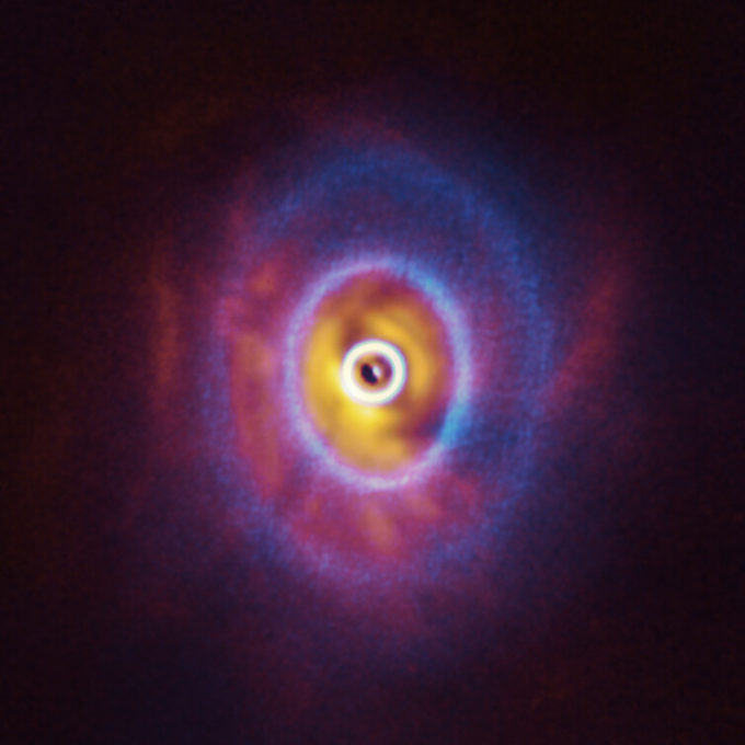 ALMA y el instrumento SPHERE del Very Large Telescope de ESO permitieron obtener estas imágenes de GW Orionis, un sistema estelar triple con peculiares características en la zona interna. A diferencia de los discos protoplanetarios planos observados alrededor de muchas estrellas, GW Orionis tiene un disco curvo, deformado por los movimientos de las tres estrellas que alberga en el centro. Esta Imagen compuesta combina las observaciones de ALMA y SPHERE realizadas por Stefan Kraus y su equipo. La imagen de ALMA (en azul) muestra la estructura en anillos, donde el anillo más céntrico (en parte visible como un ovoide en el centro de la imagen) está separado del resto del disco. Las observaciones de SPHERE (en rojo y naranja) permitieron a los astrónomos detectar por primera vez la sombra del anillo interno sobre el resto del disco y, de esa forma, reconstituir su forma curva. Créditos: ALMA (ESO/NAOJ/NRAO), ESO/Exeter/Kraus et al.