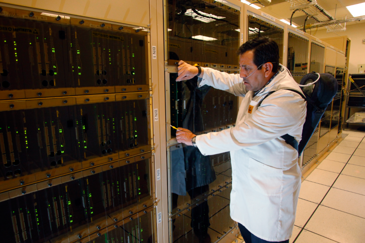 El técnico Juan Carlos Gatica revisa instrumentos electrónicos del correlacionador de ALMA. Para llevar a cabo la inspección en las instalaciones, ubicadas a gran altitud, Juan Carlos usa suministro de oxígeno. ALMA obtuvo recientemente el visto bueno para realizar importantes mejoras en el correlacionador –el cerebro– del telescopio. También se aprobaron actualizaciones para el sistema de transmisión digital, la autopista de transmisión de datos del observatorio. Créditos: Carlos Padilla, NRAO/AUI/NSF