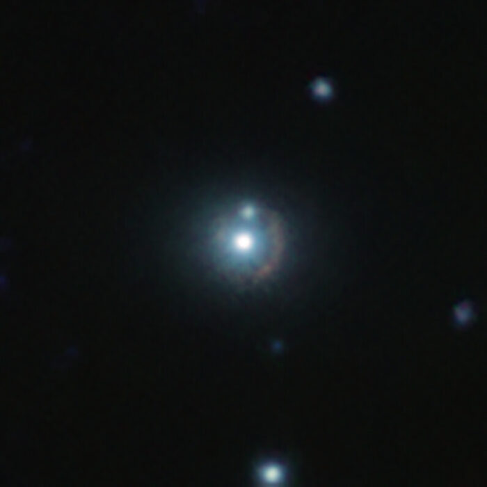 Esta imagen infrarroja muestra la galaxia distante 9io9, vista aquí como un arco rojizo curvado alrededor de una galaxia cercana brillante. Esta galaxia cercana actúa como una lente gravitacional: su masa curva el espacio-tiempo a su alrededor, doblando los rayos de luz provenientes de 9io9, al fondo, de ahí su forma distorsionada. Esta vista a color es el resultado de combinar imágenes infrarrojas tomadas con el telescopio VISTA (Visible and Infrared Survey Telescope for Astronomy, telescopio de sondeo en el visible y el infrarrojo para astronomía) de ESO, en Chile, y el CFHT (Canada France Hawaii Telescope, telescopio Canadá, Francia y Hawái), en Estados Unidos. Crédito: ESO/J. Geach et al.